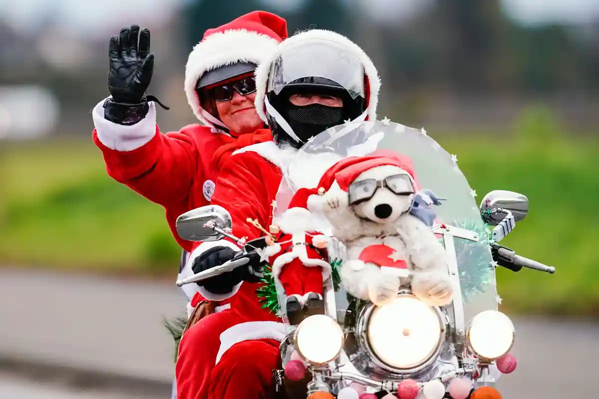 Санта на мотоцикле:Мотоциклисты в костюмах Санты едут по проселочной дороге и машут руками.