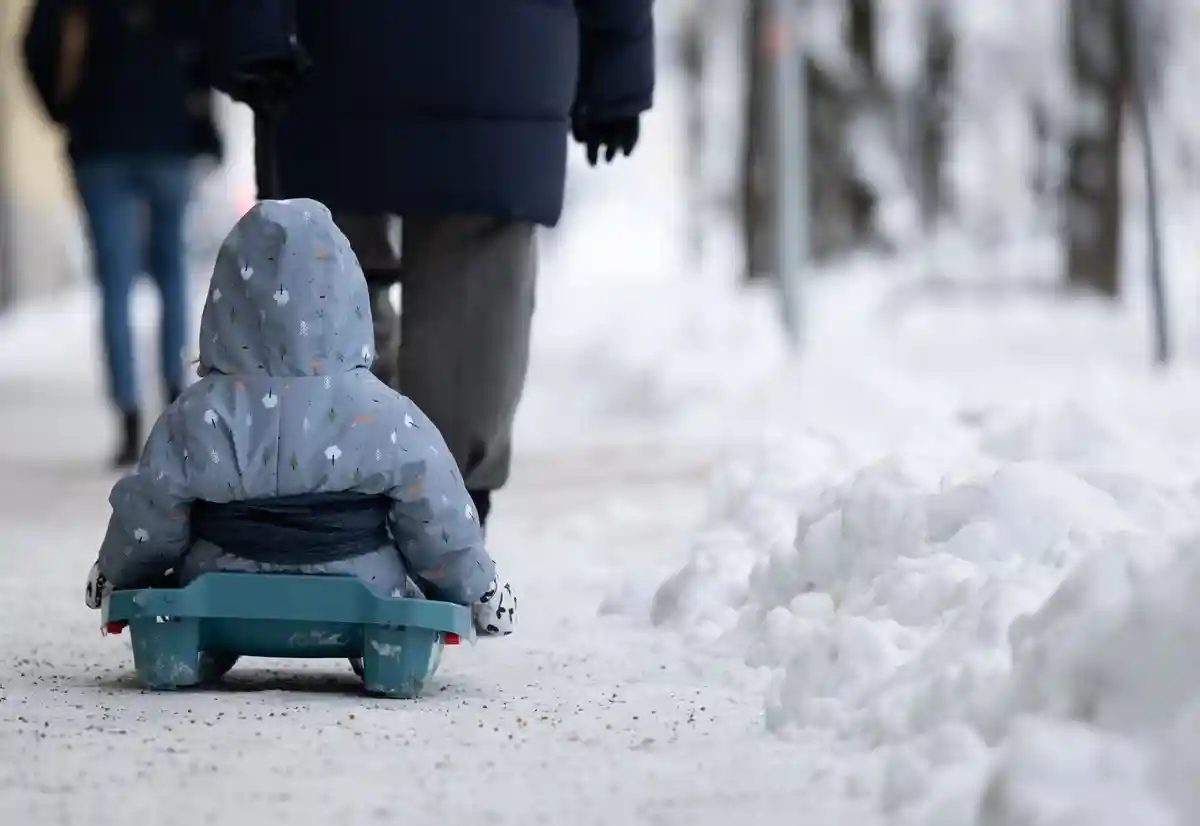 Зимняя погода в Германии - Мюнхен:Женщина везет ребенка на санках по тротуару.
